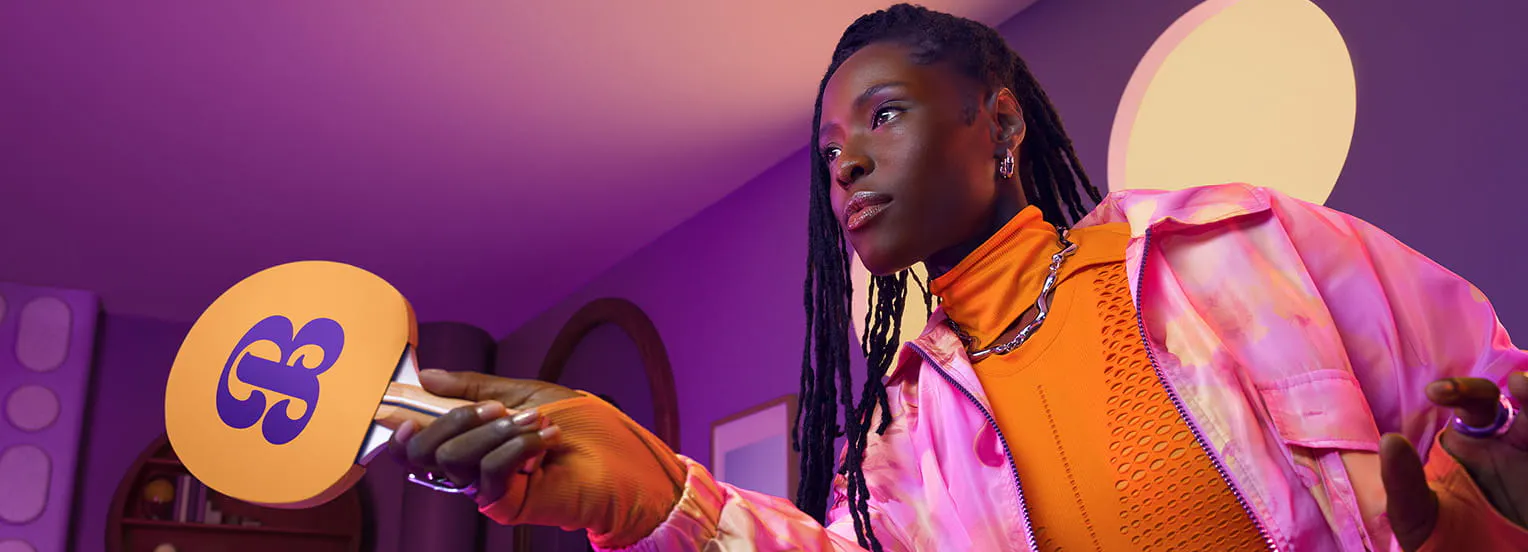 Woman in an orange shirt and jacket holding a yellow ping pong paddle