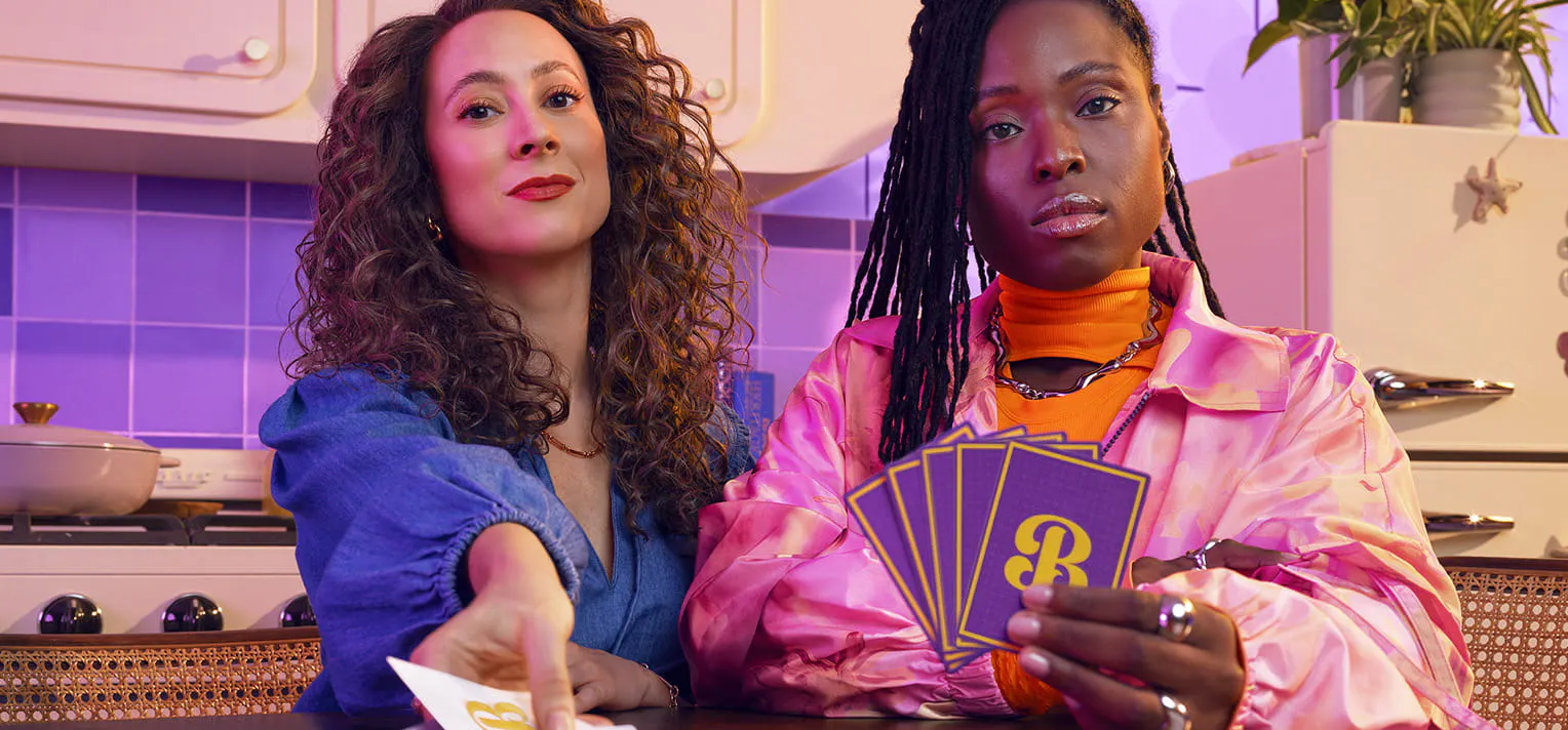 Woman holding playing cards with the letter B on them while another woman places a card on the table