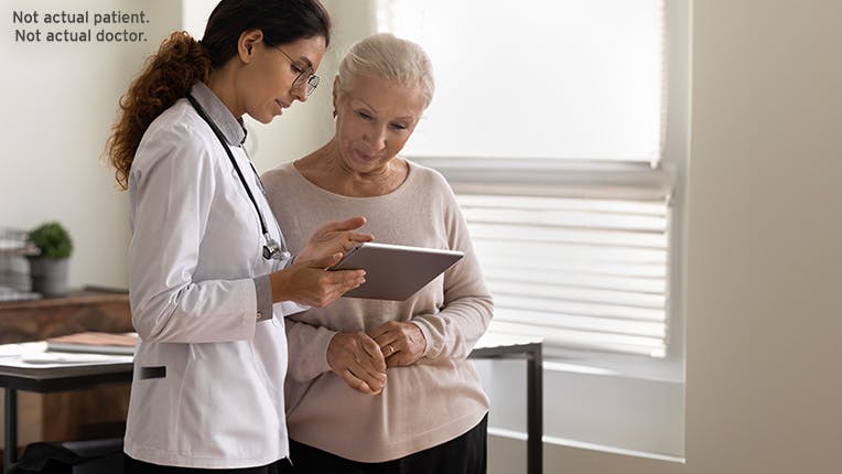  A female acting as a doctor is holding a tablet while talking to a woman