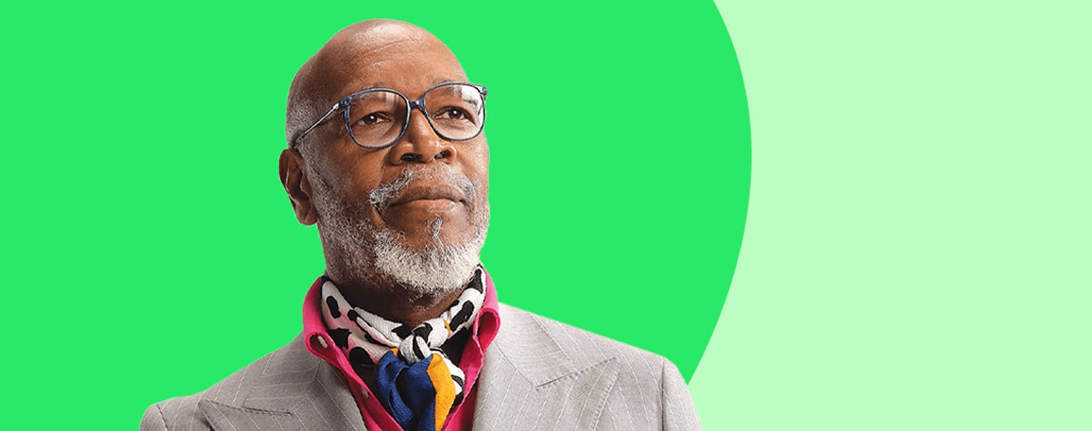 A black male wearing glasses, a grey suit and pink shirt with a colorful ascot.