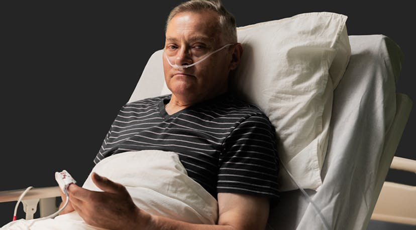 Older man with finger pulse oximeter and oxygen tubes in nose sitting in hospital bed
