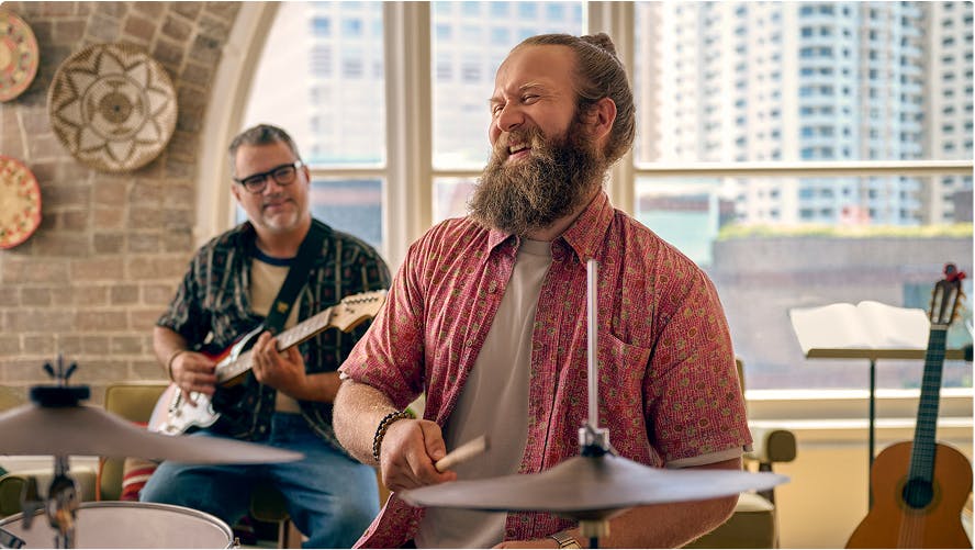Smiling man playing drums with another man playing guitar