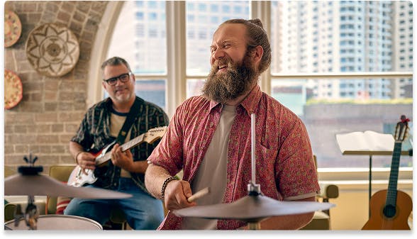 Smiling man playing drums with another man playing guitar