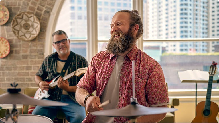 Smiling man playing drums with another man playing guitar