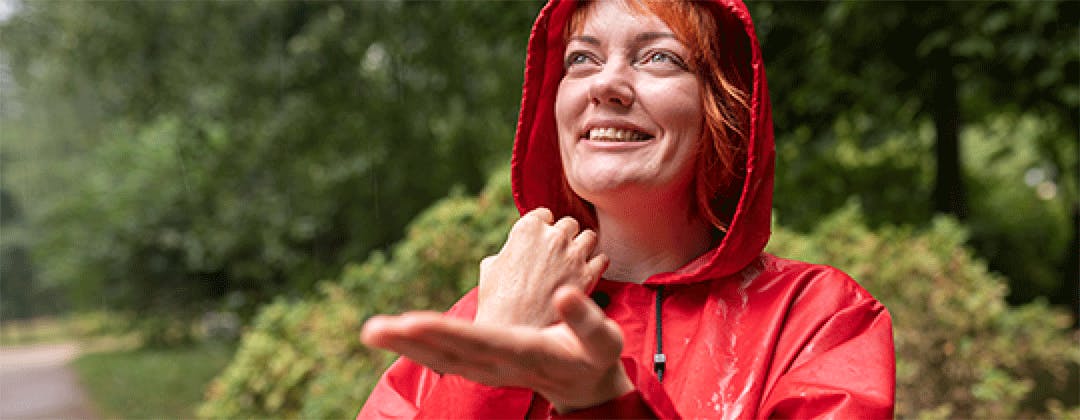 A woman with a red rain coat standing in a park in the rain smiling up at the sky catching rain drops