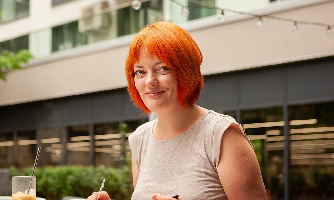 Caucasian women with short orange hair with a fridge, sits at a table having a beverage, writing, looking into the camera