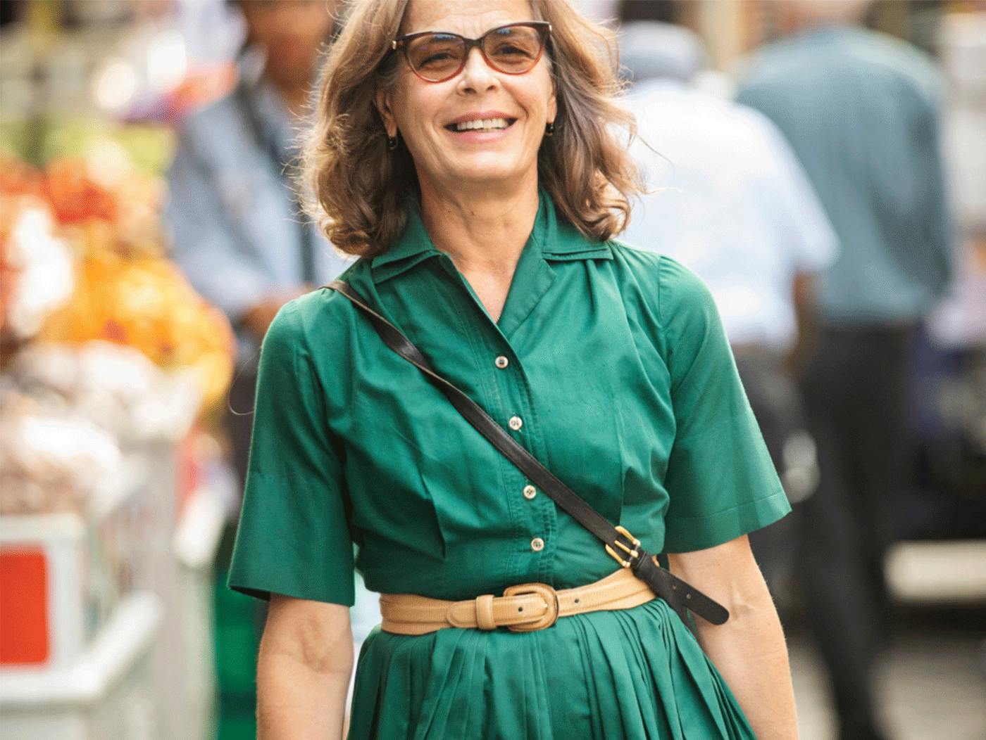 An elder caucasian woman with a jade green dress smiling while walking through a street market on a sunny day