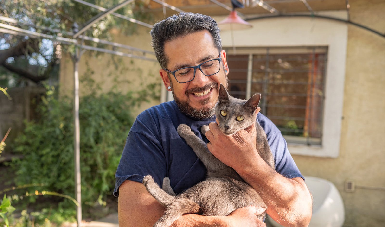 A bearded caucasian man with dark hair and glasses holding a grey cat and smiling in his backyard.  “I regularly discuss quality of life, and where I want my wellbeing to be, with my doctor.” “I regularly discuss quality of life, and where I want my wellbeing to be, with my doctor.” Sam, living with HIV