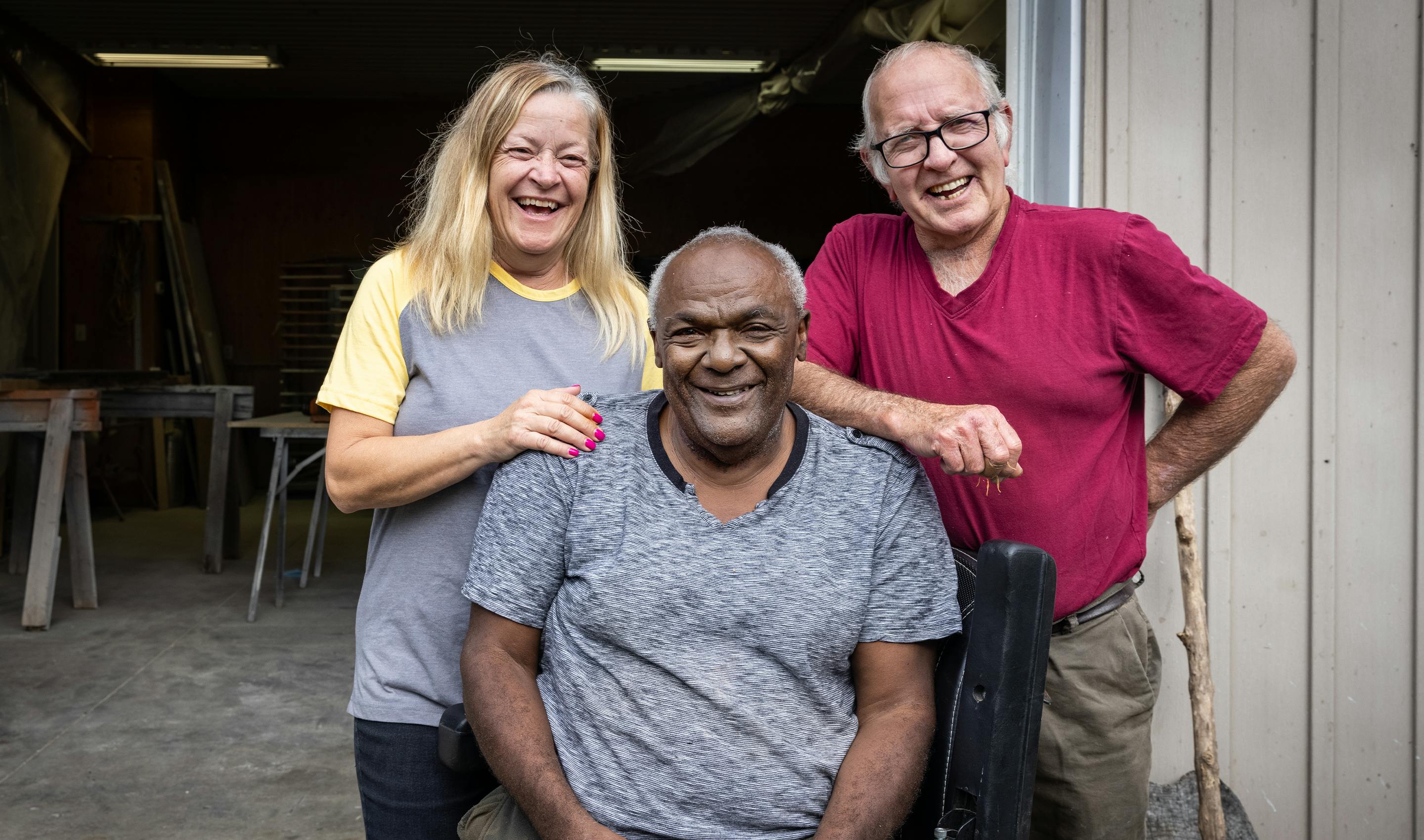 An older African man seated in a wheelchair smiling at the camera flanked by two friends standing behind him.