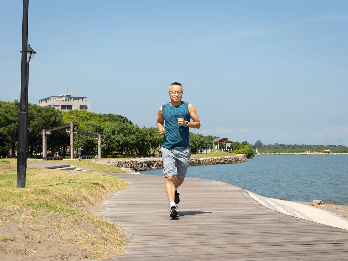 A middle aged asian man running along a wooden boardwalk by the water in the sun