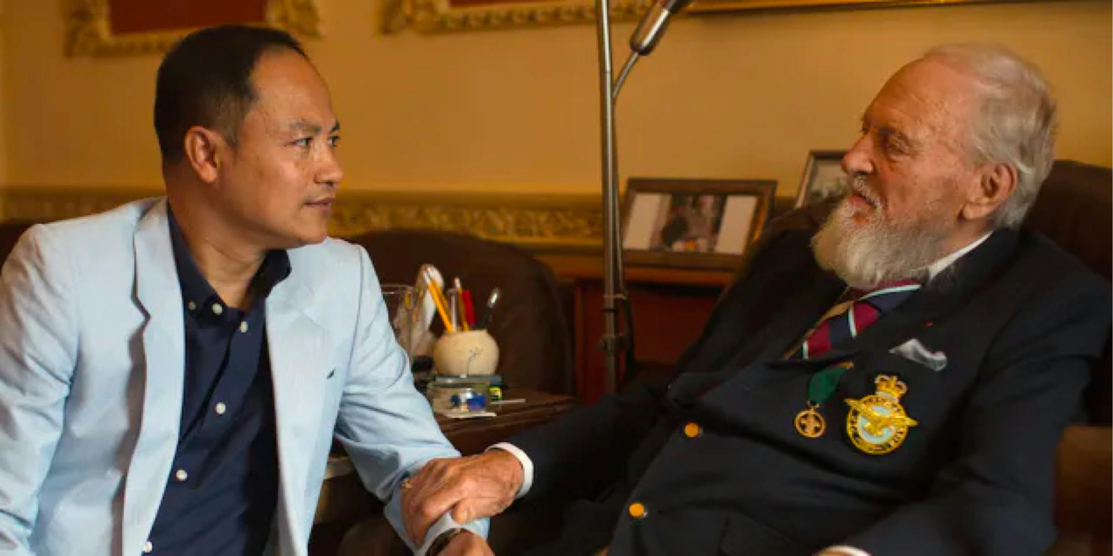 An older man sits with another man, resting his hand on the other’s arm