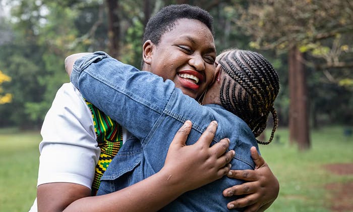 Two people embracing outdoors, one in a denim jacket with braided hair and the other in a patterned white shirt, with trees and greenery in the background.