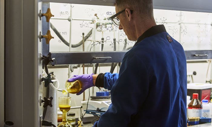 Person in blue lab coat and purple gloves pouring yellow liquid in a lab, with equipment and chemical formulas in the background.