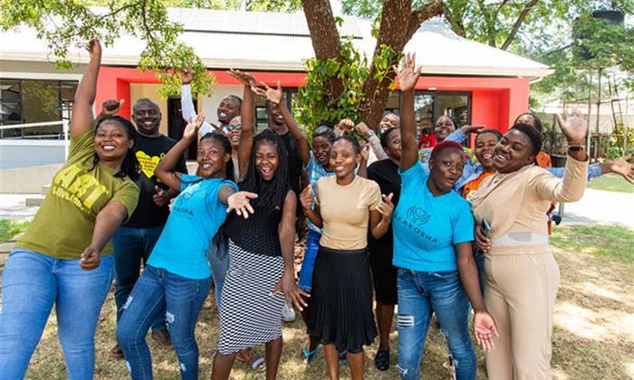 Group of people standing on grass in front of a red and white building, arms raised in a celebratory pose.