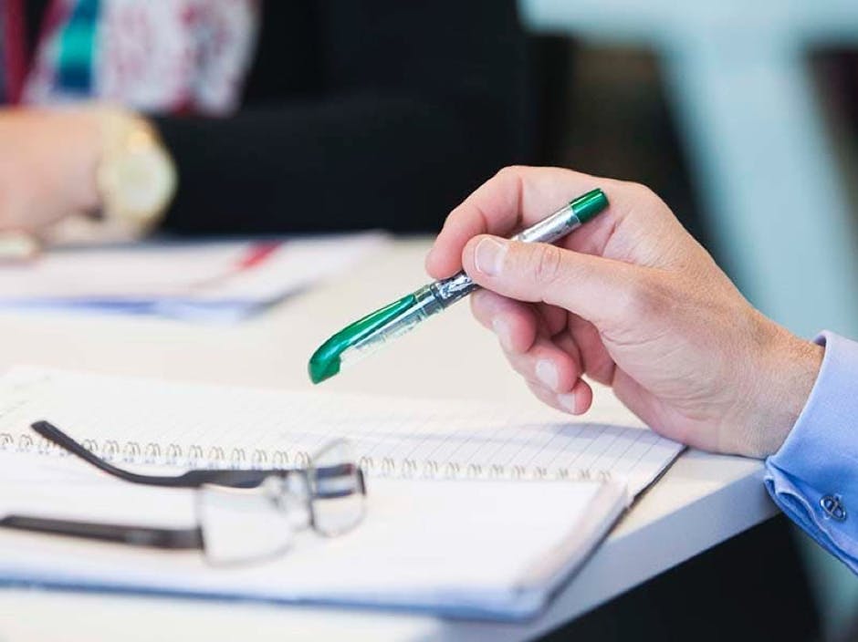 Close up of hands on a table working, with a pair of glasses sitting on it and a hand holding a pen